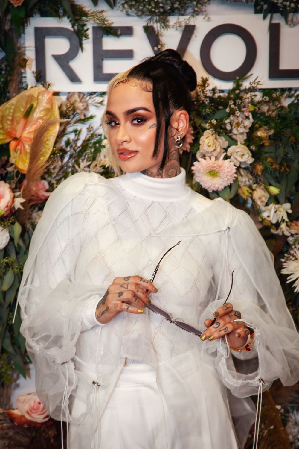 Woman in white sheer outfit with tattoos poses at Revolve event amid floral backdrop.