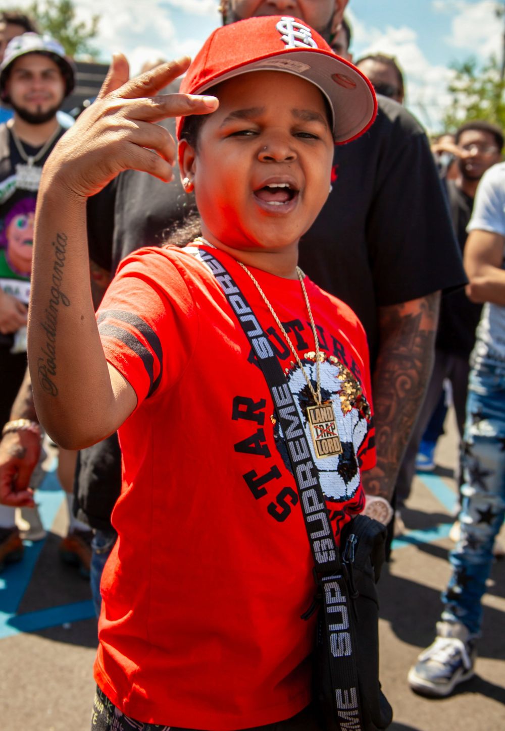 A young boy in a red Supreme shirt and St. Louis Cardinals cap poses confidently at an outdoor event.