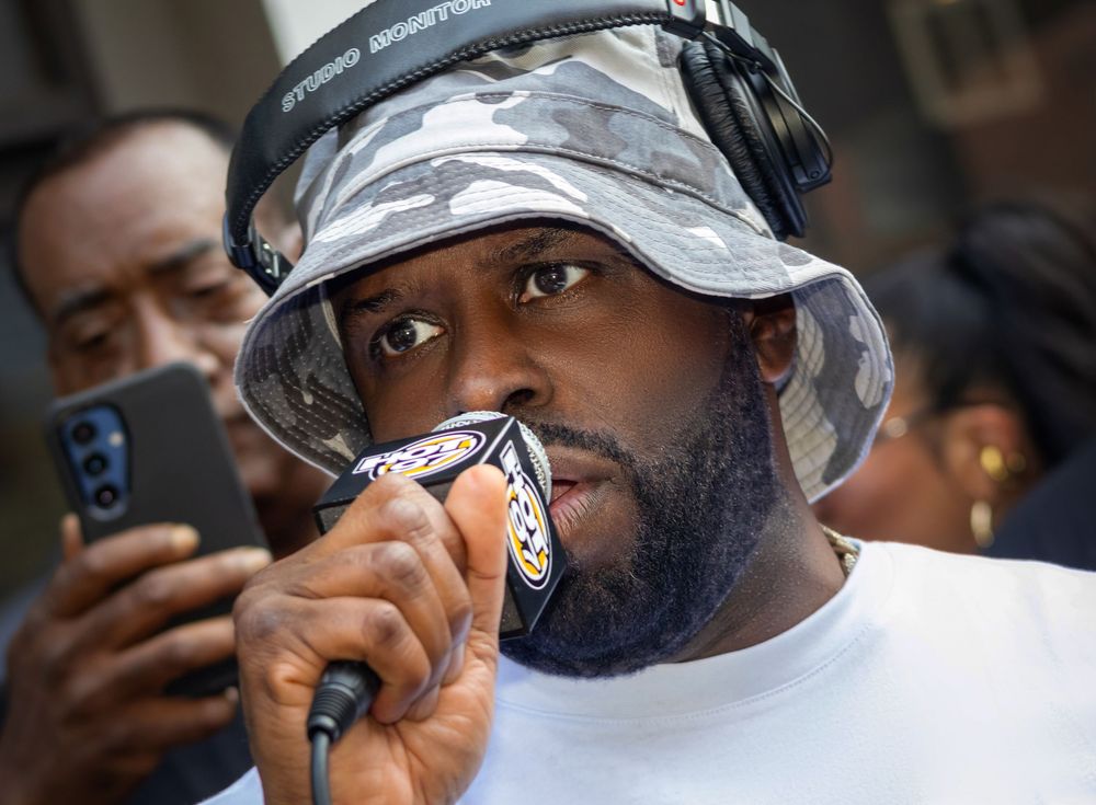 DJ Funk Flex speaking into microphone at outdoor event, wearing camo bucket hat and headphones.