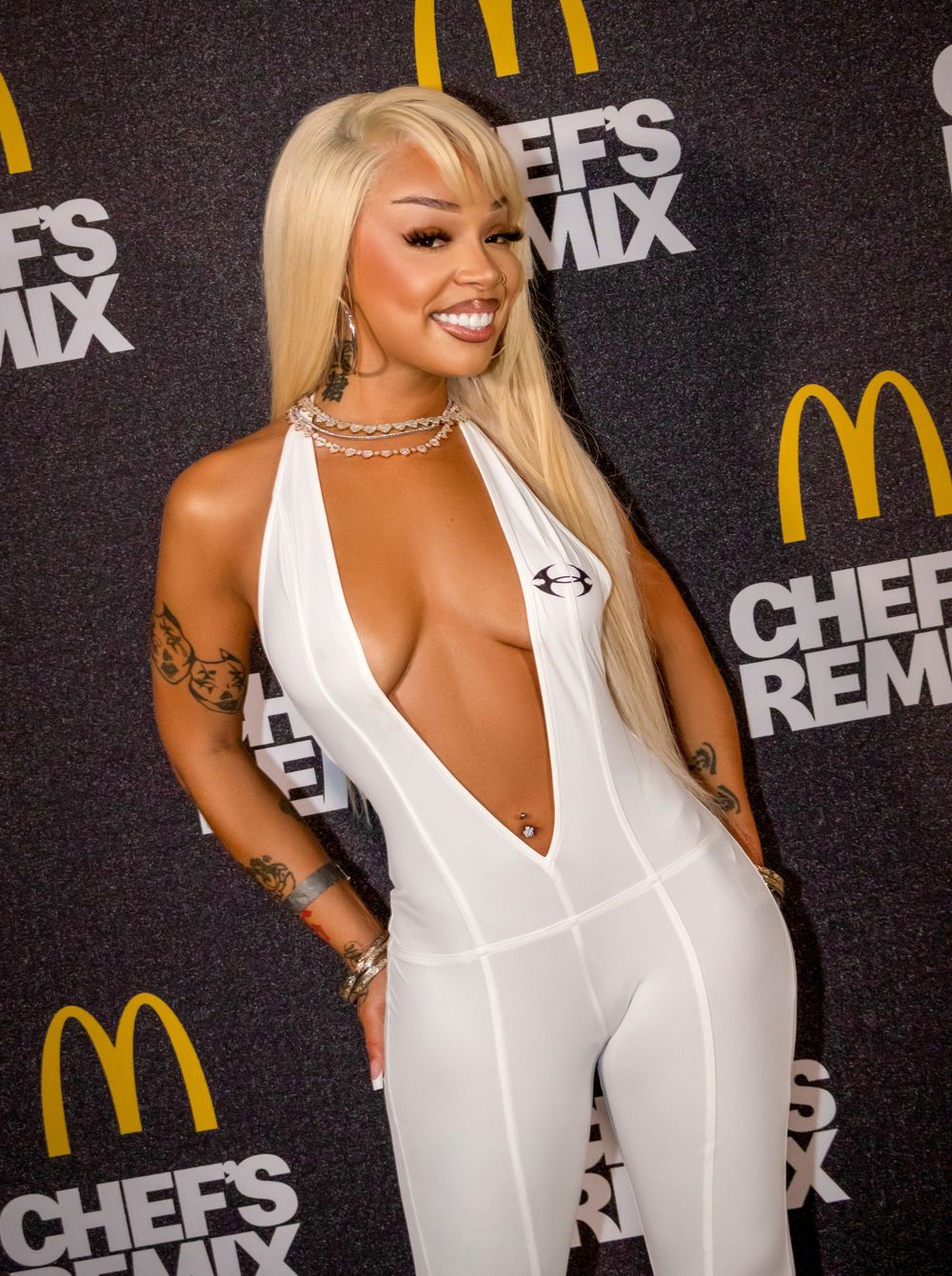 Honey Bxby in white deep-V jumpsuit with blonde hair posing at McDonald's Chef's Remix event on black carpet.