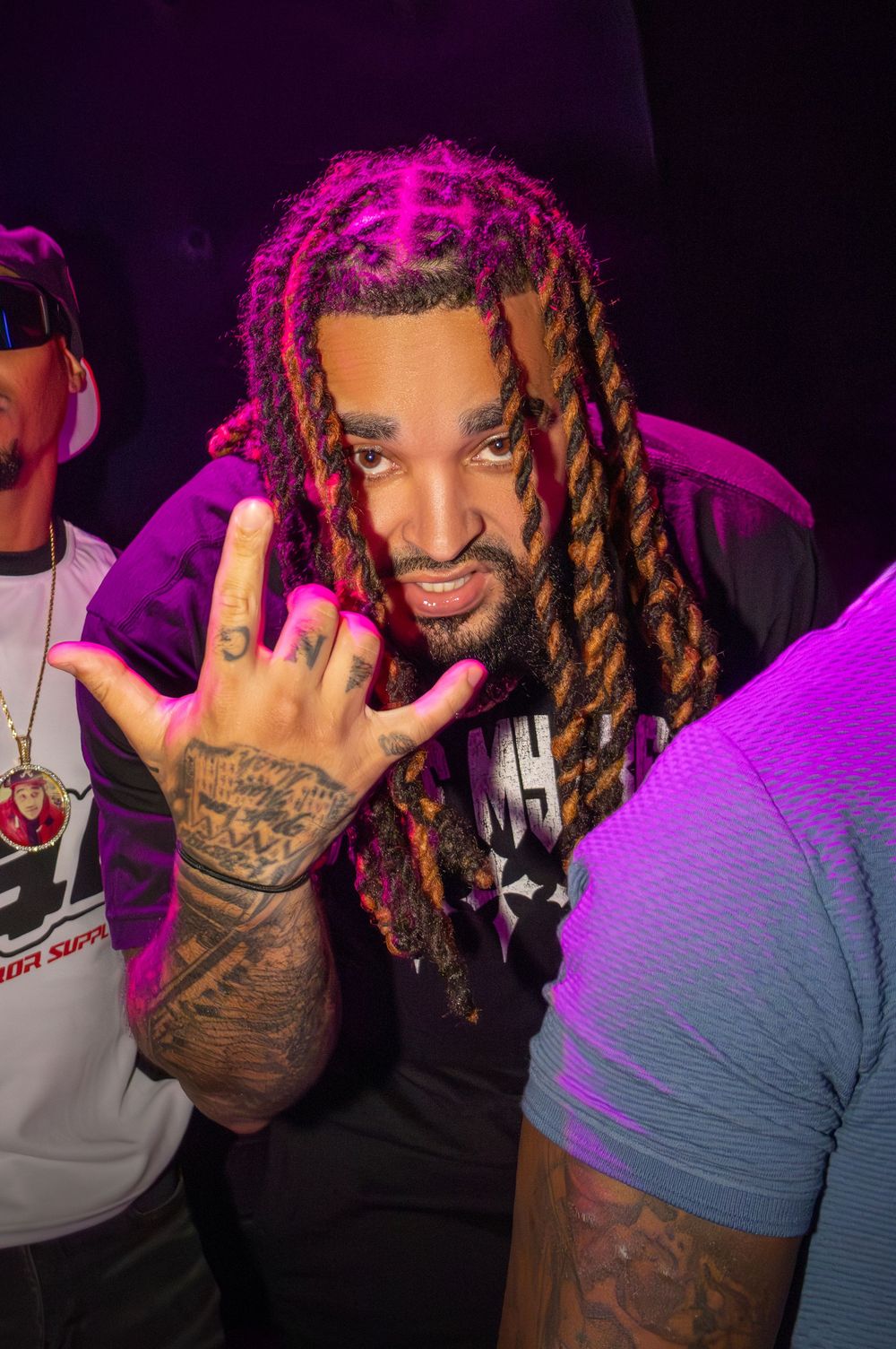 Albee AL with pink dreadlocks and hand tattoos flashing signs under purple club lighting.