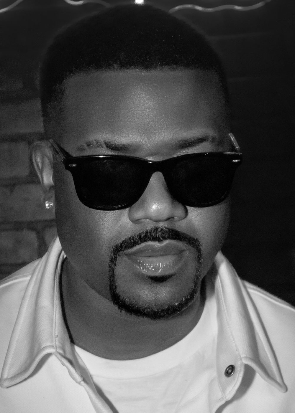 Ray J wearing black sunglasses with goatee in black and white portrait photo.