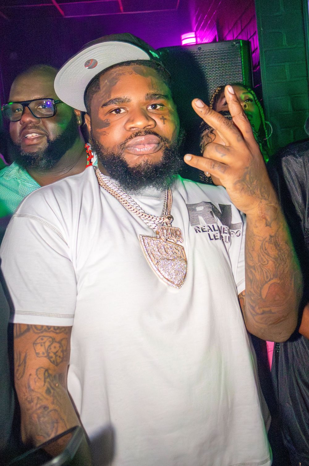 Fatboy SSE with a beard wearing a white t-shirt and cap poses with chain jewelry at a nightclub event.