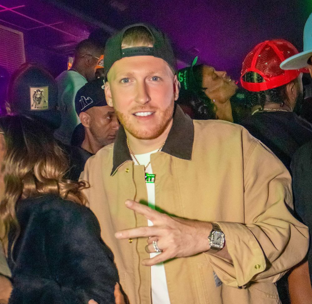 DJ Drewski wearing a backwards green cap and tan jacket poses at a nightclub party, flashing a hand sign.
