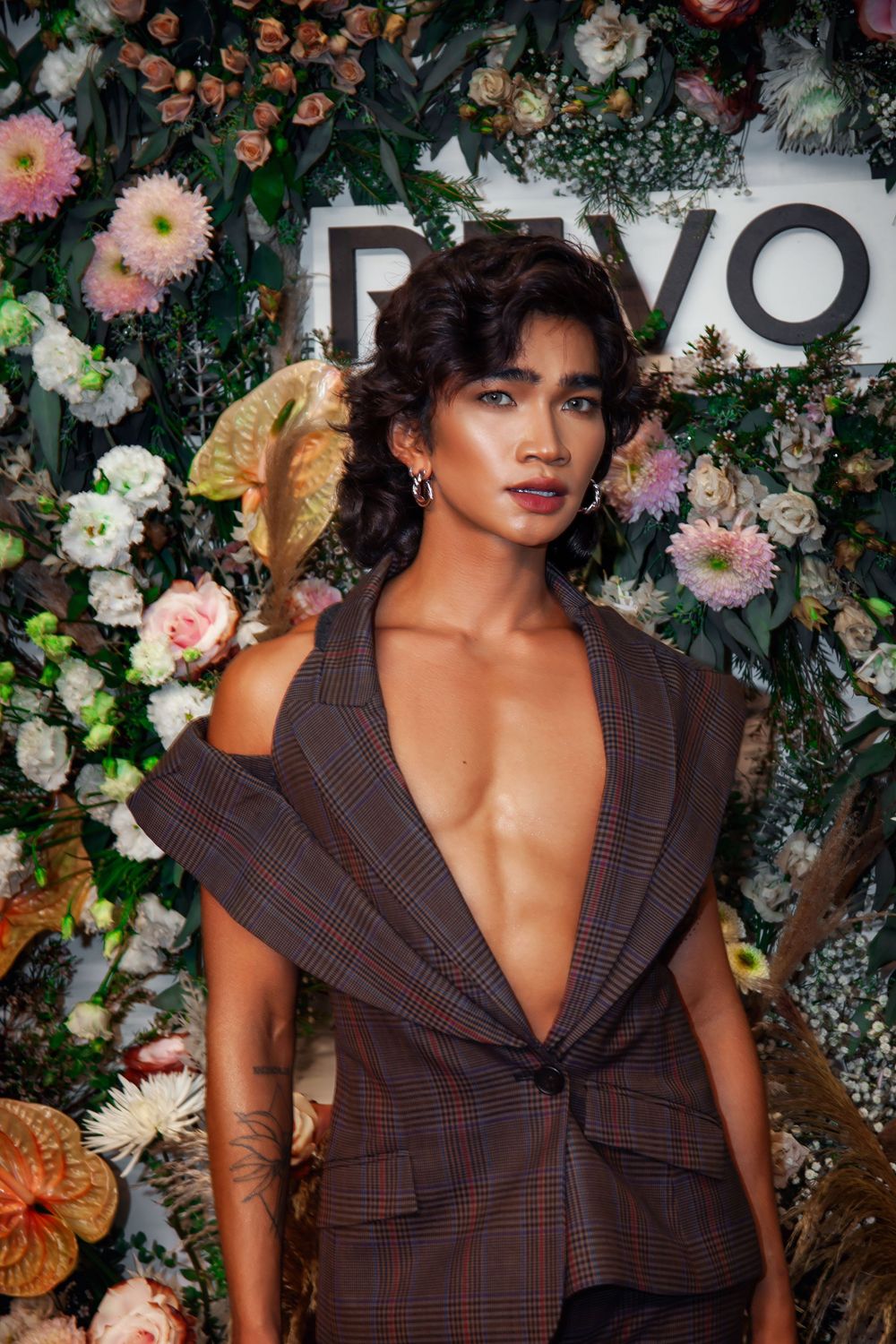 A woman in a deep V-neck plaid blazer poses in front of a floral wall at a Revolve event.