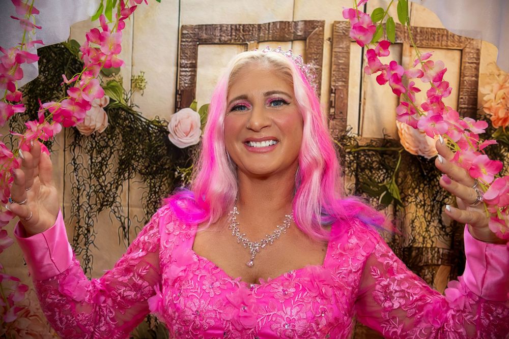 Woman in pink wig and lace dress surrounded by pink orchid flowers, smiling joyfully at a floral decorated event.