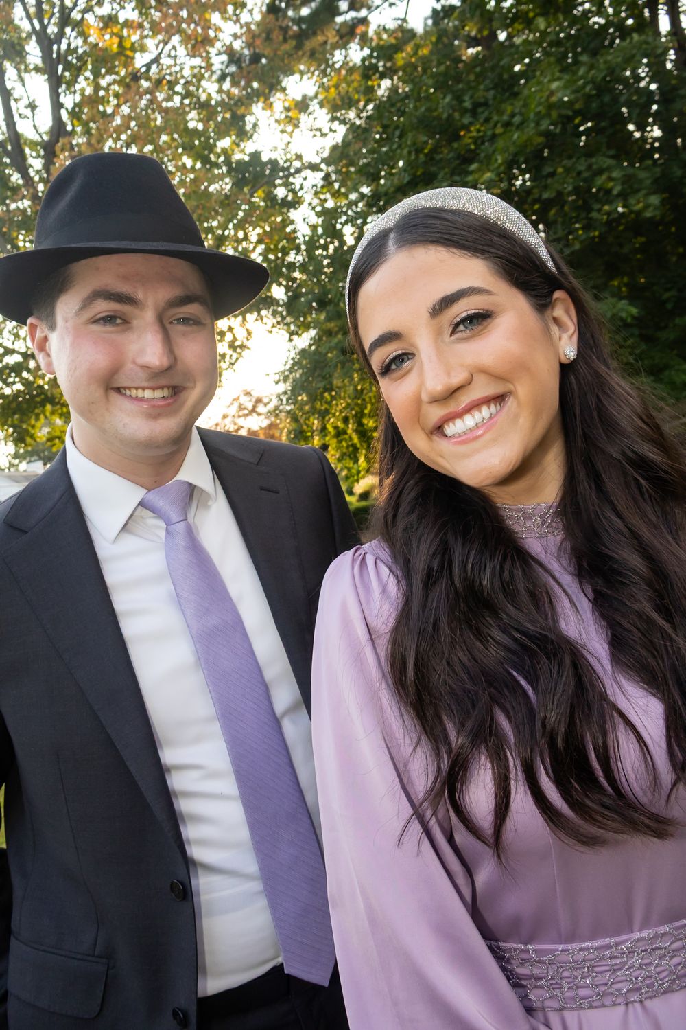 Smiling couple in formal attire, man in black suit and hat, woman in lavender dress, outdoors with trees.