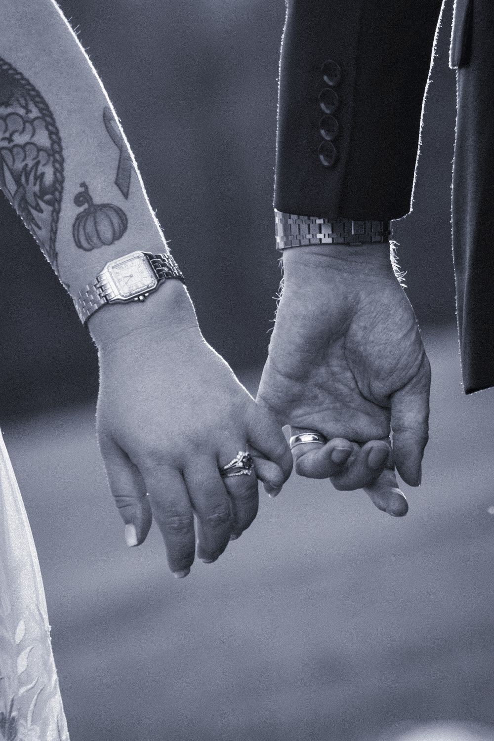 Black and white photo of newlyweds holding hands, showing wedding rings, a tattoo, and elegant watches.