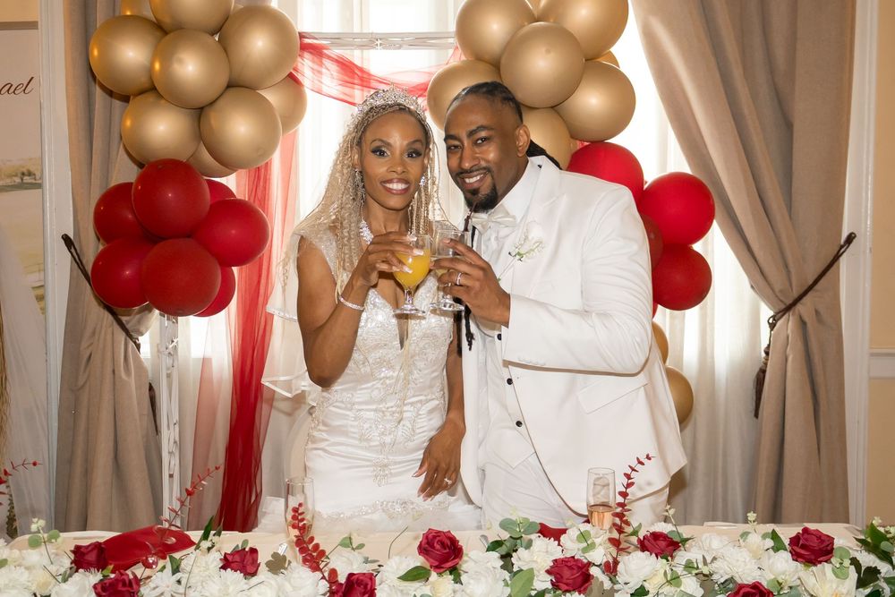 Bride and groom toasting champagne at wedding reception with red and gold balloon arch backdrop.