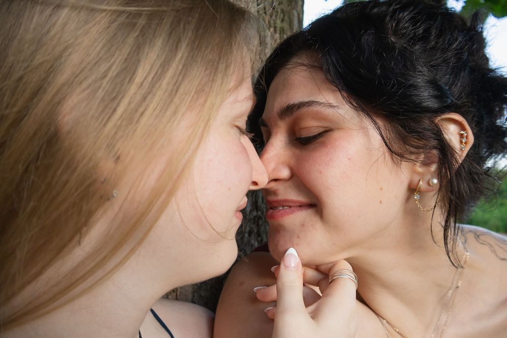 Two young women sharing a close intimate moment outdoors near a tree, one blonde and one brunette.