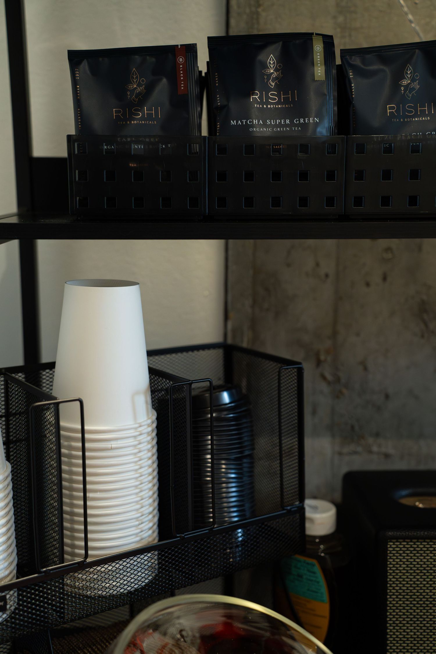 Organized shelf with Rishi tea packages and white disposable cups in metal storage basket.
