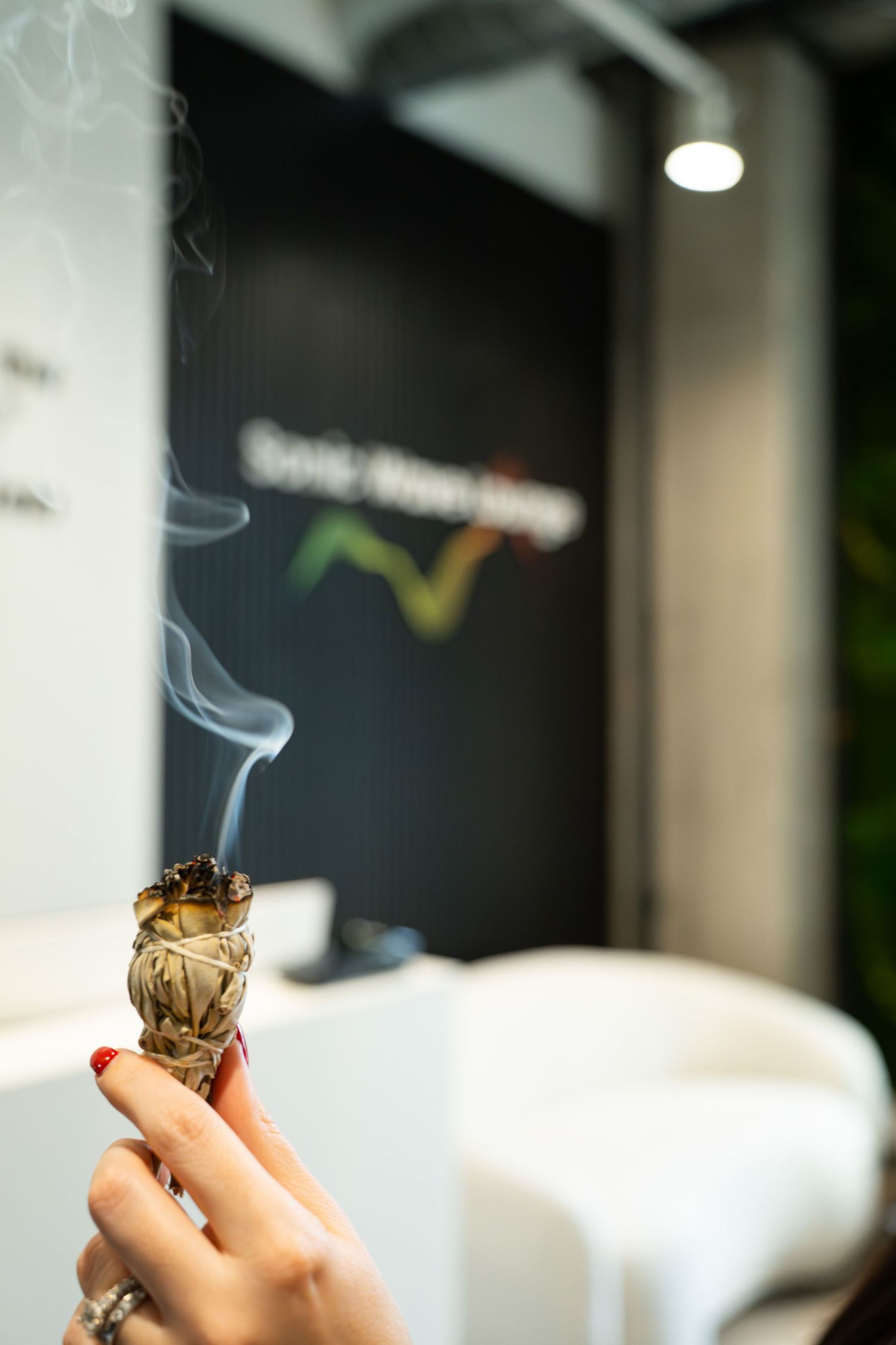 Hand holding burning sage or smudge stick against blurred dark background with logo.