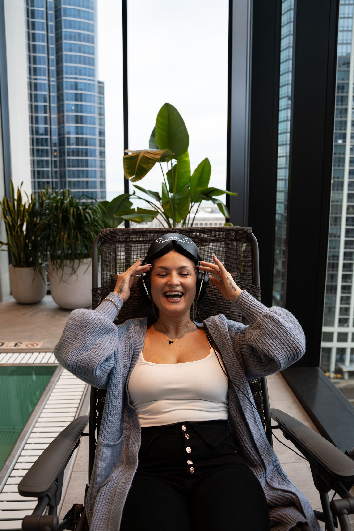 Someone enjoying music in a lounge chair by floor-to-ceiling windows overlooking city buildings.