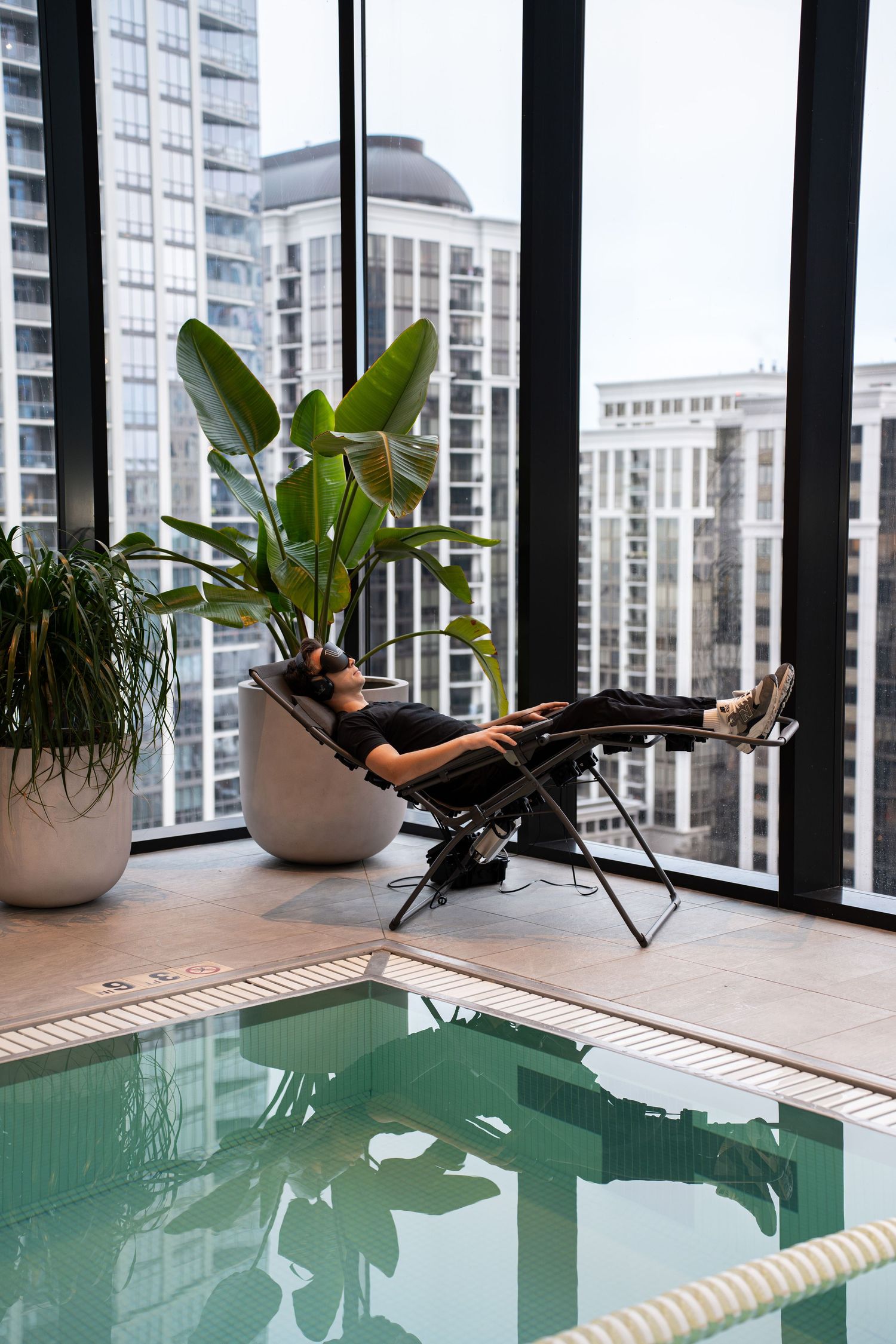 Modern high-rise apartment with indoor pool and lounge chair next to large windows.