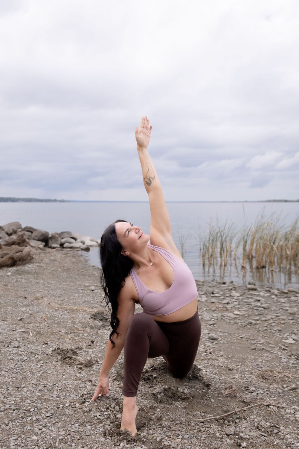 Yoga stretch performed on rocky beach with lake and cloudy sky in background.