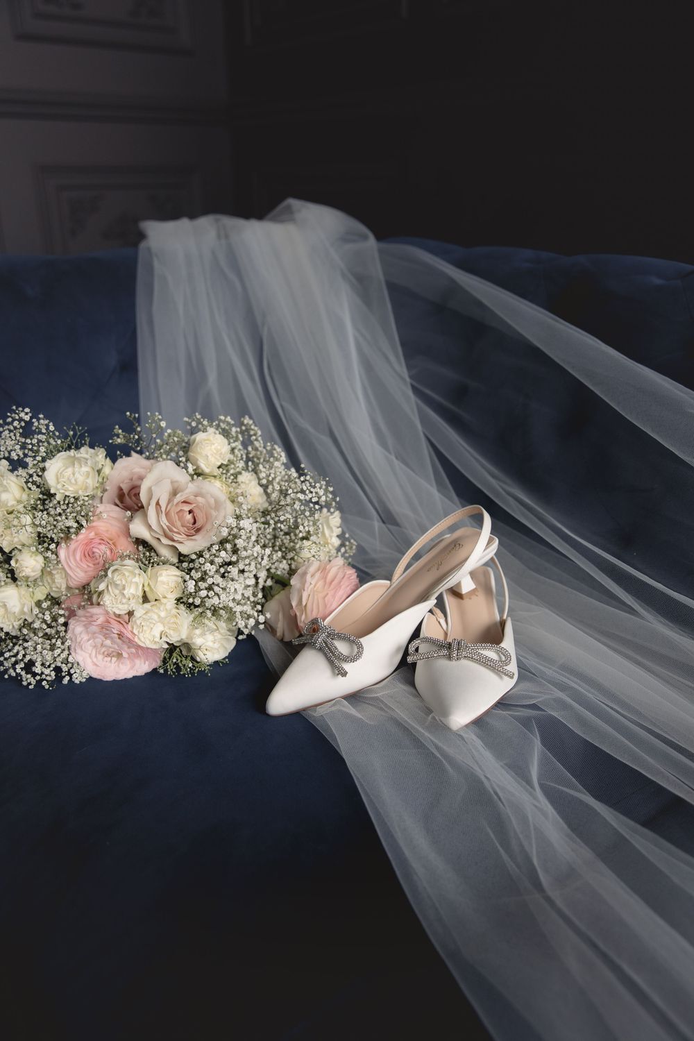 Elegant white bridal shoes with bows displayed alongside a soft pink and white bouquet on flowing tulle veil.