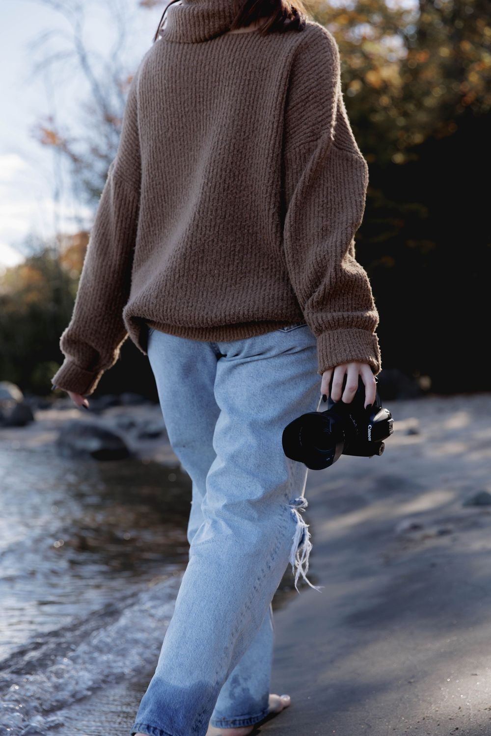 Person wearing brown sweater and light blue jeans walks along rocky shoreline at sunset.