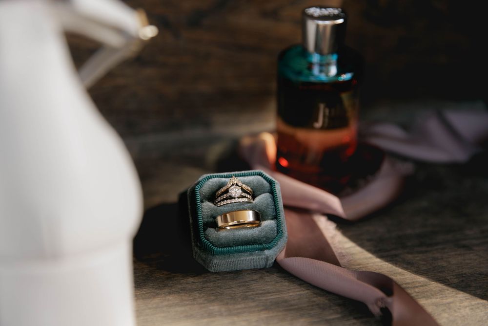 Two engagement rings in teal velvet ring boxes sit on a dark wooden surface next to a bottle of perfume.