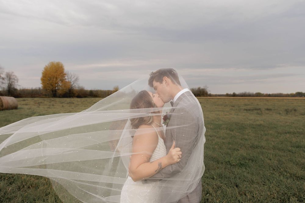 A romantic wedding couple embraces in a grassy field as a long veil flows in the autumn breeze.