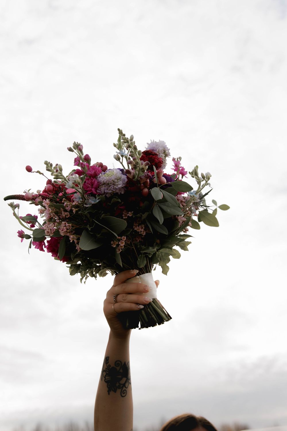 Hand holding a bouquet of pink flowers and eucalyptus leaves against a cloudy sky.