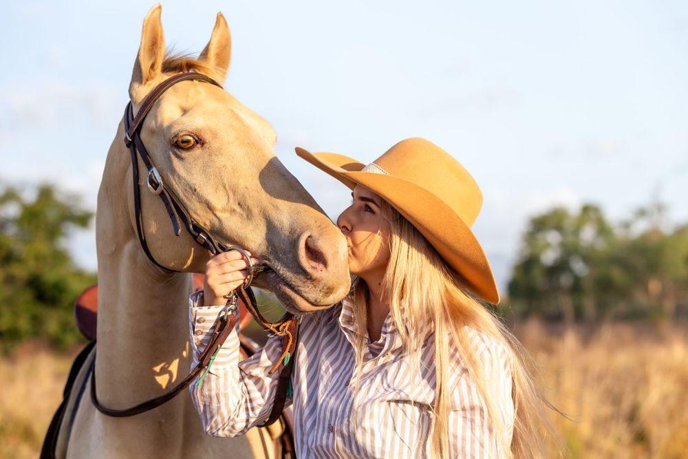 Person in brown hat lovingly embraces palomino horse in golden sunlit field during sunset.