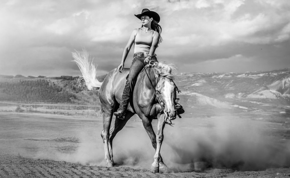 A dramatic black and white western scene shows a rider on horseback kicking up dust against a mountain landscape.