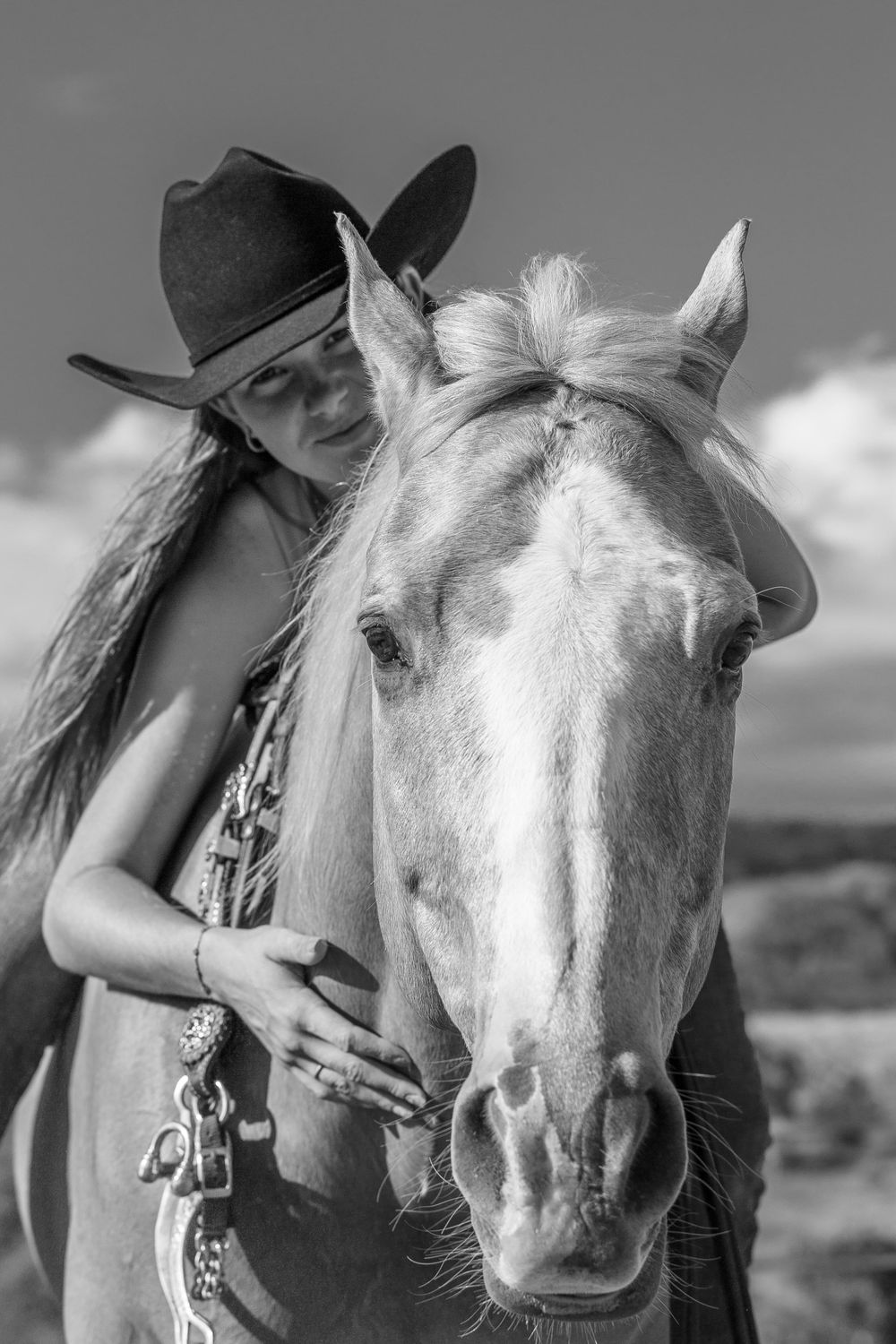A close-up black and white portrait showing a rider in a cowboy hat alongside their white horse against a cloudy sky.