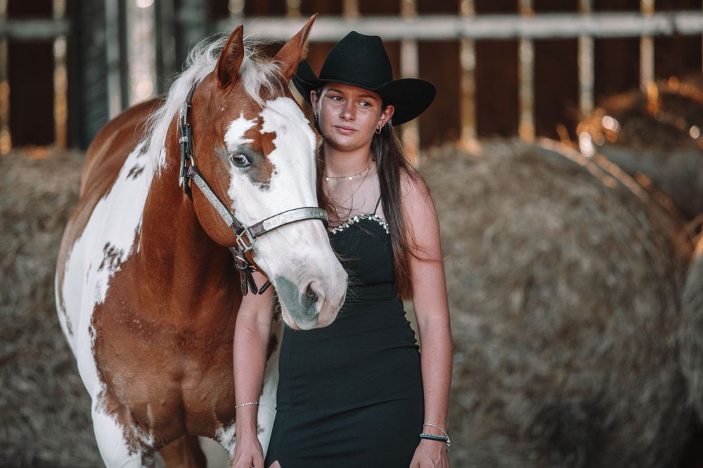 Brown and white paint horse stands next to someone wearing a black dress and cowboy hat in a rustic barn setting.