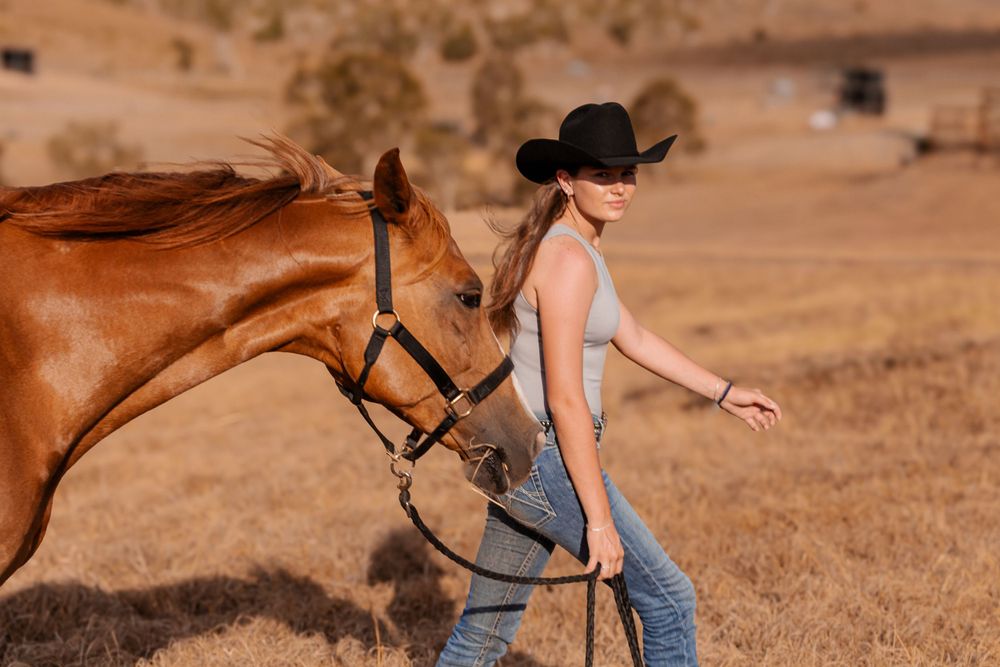 A chestnut horse follows its rider wearing a black cowboy hat across a sunlit pasture.