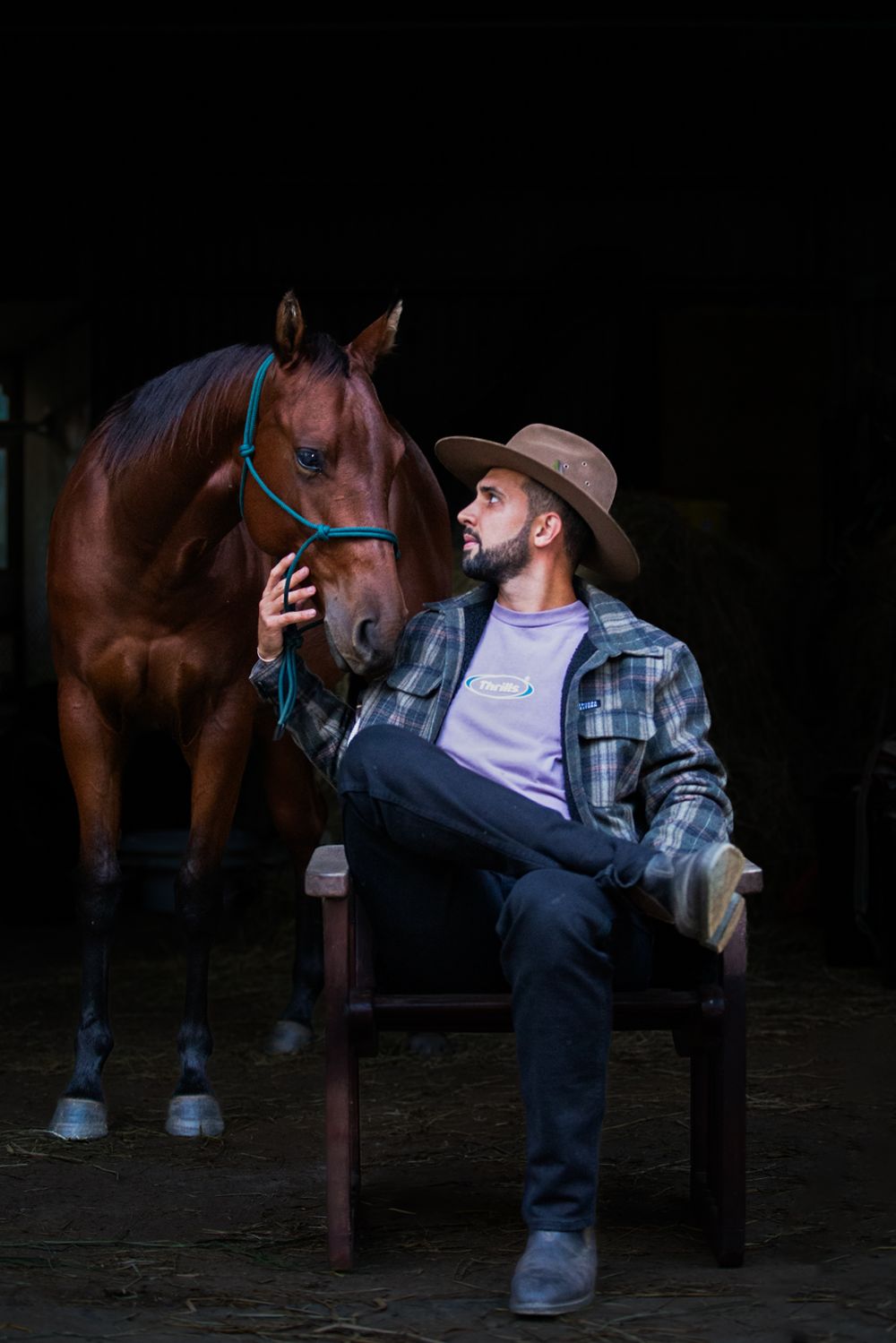 A rancher in plaid shirt and cowboy hat sits casually on a fence post while bonding with his chestnut horse in a dark barn.