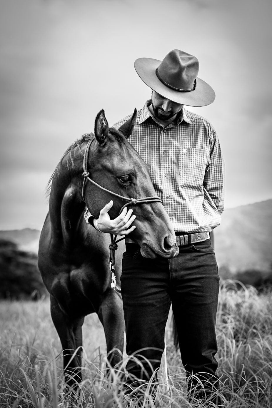 Black and white portrait of a cowboy in a hat and plaid shirt standing with a dark horse in tall grass.