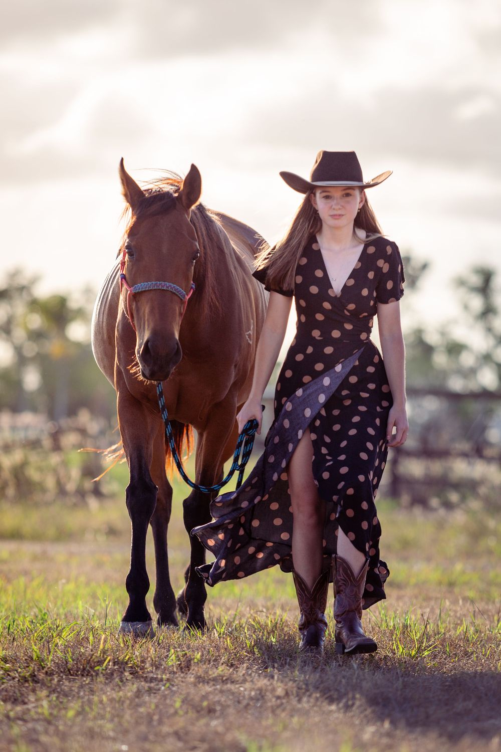 A person in a polka dot dress and cowboy hat walks alongside a brown horse in a sunlit field at dusk.
