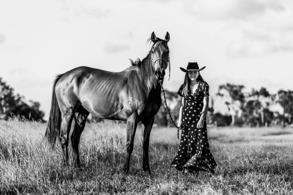 Black and white photo of a tall horse and person in polka dot dress standing in a grassy field.