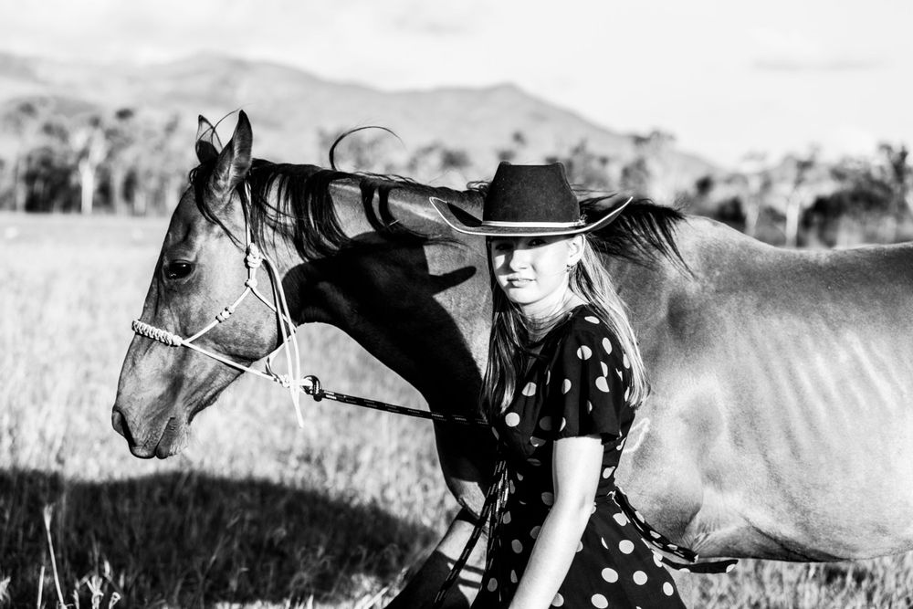 Black and white photo of a person in a cowboy hat and dress standing next to a horse outdoors.