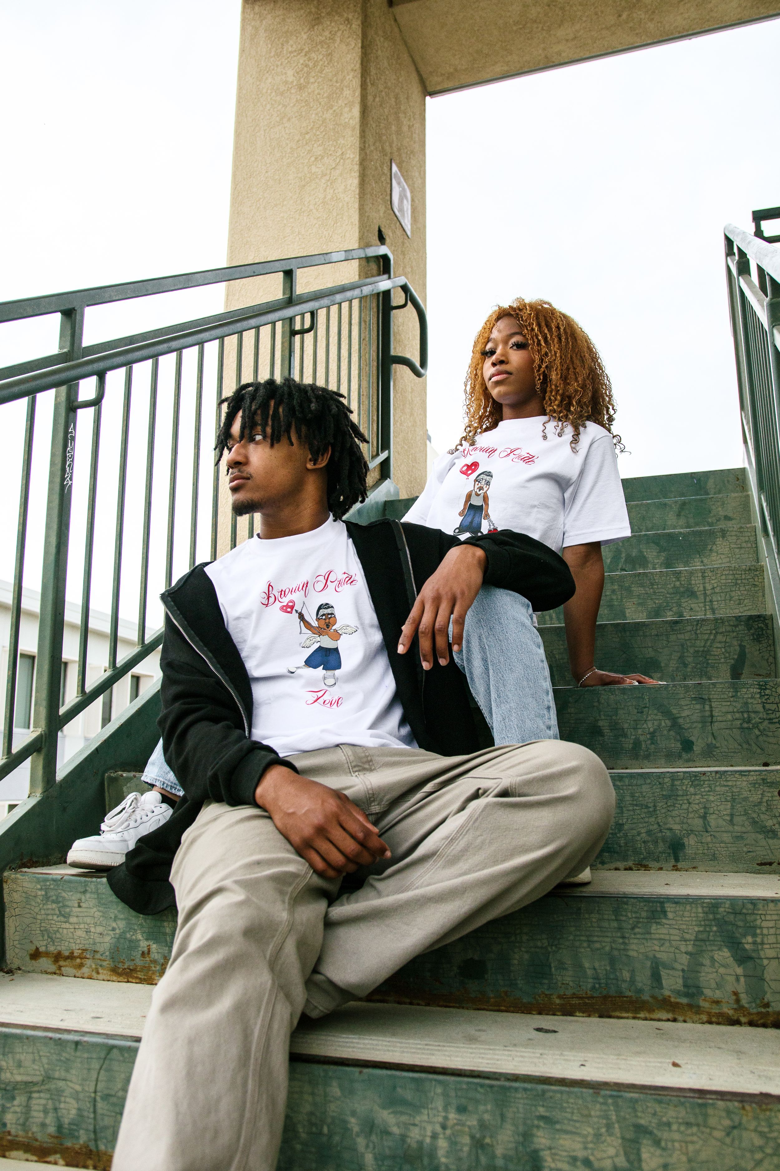 Two young adults wearing matching white Brown Pride graphic tees on outdoor stadium steps.