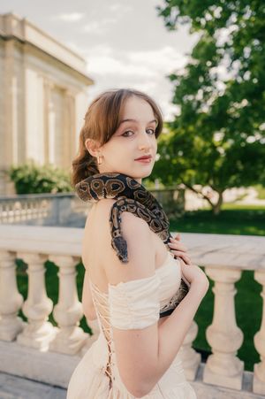 Woman in white corset dress poses on classical balcony with a large snake draped around her shoulders.