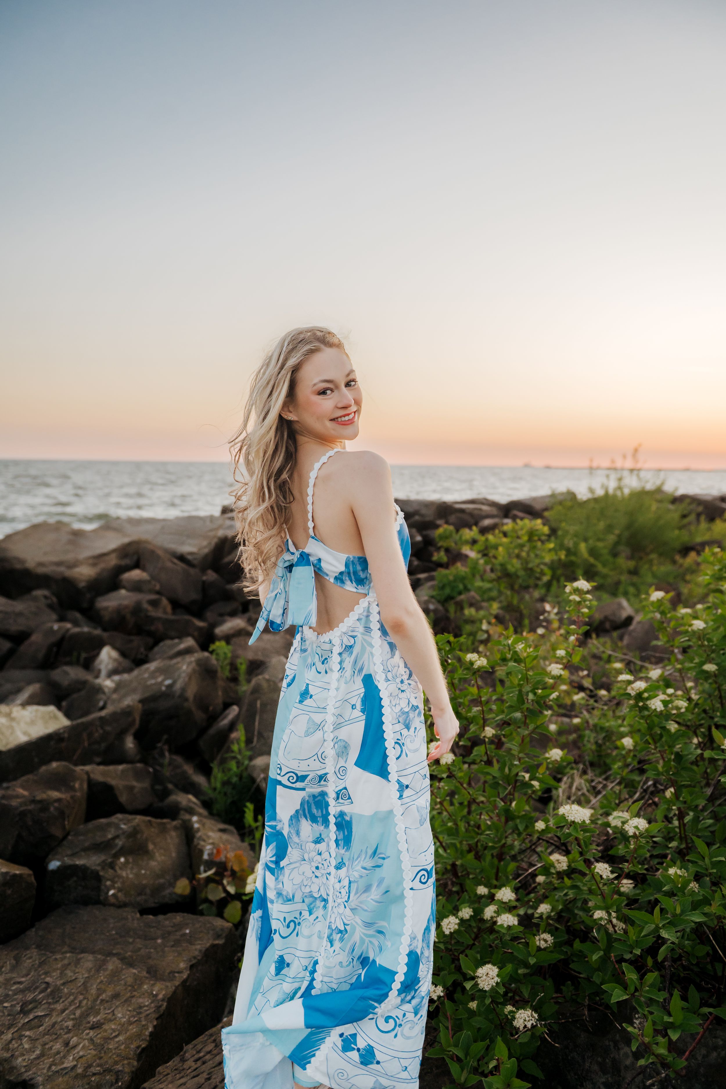 A woman in a blue and white floral sundress poses at sunset near rocky coastline with blooming wildflowers.
