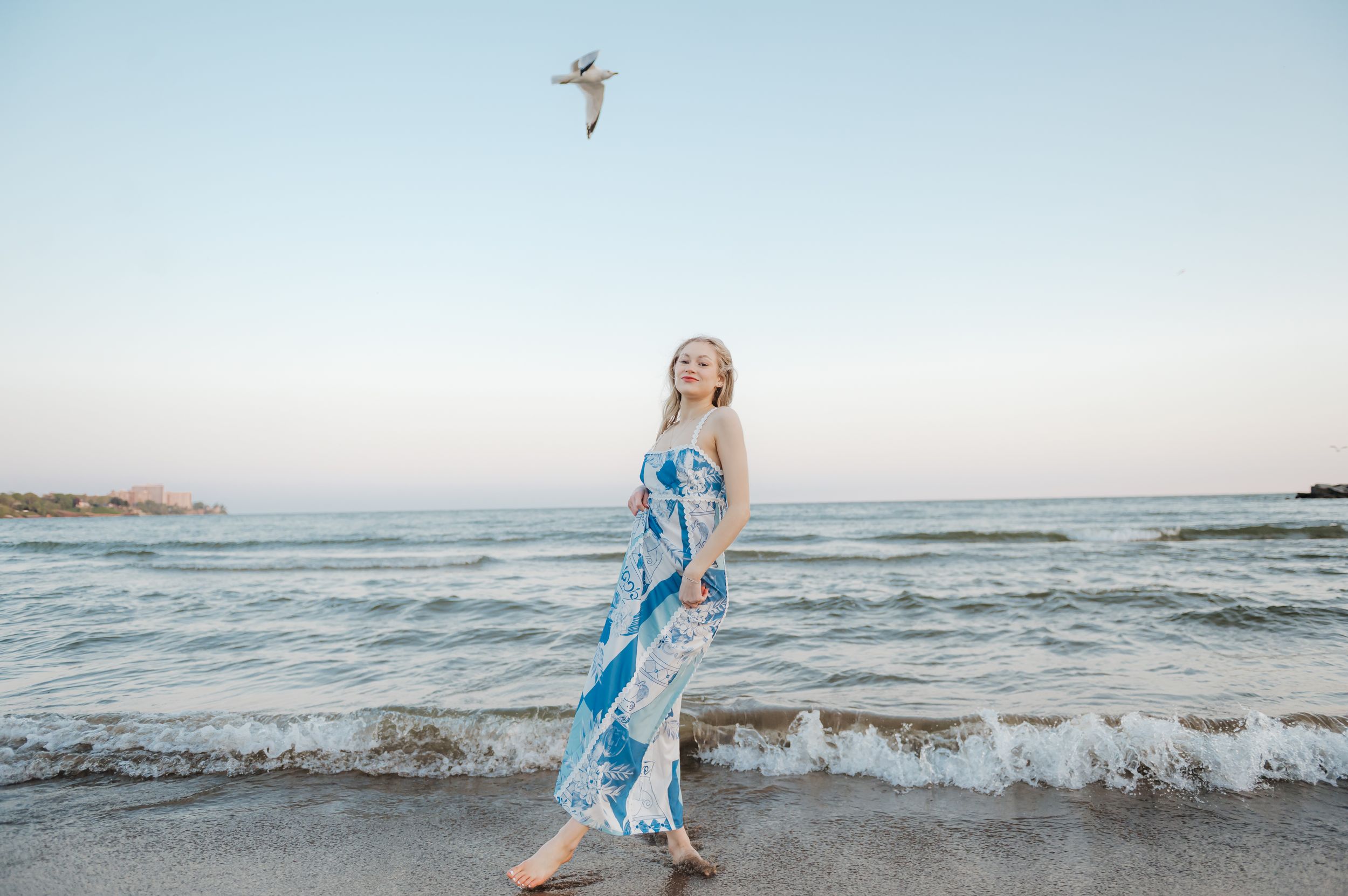 A person in a flowing blue dress stands on the beach at sunset while a seabird soars overhead.