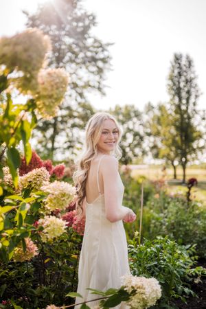 A dreamy outdoor garden photoshoot featuring someone in a flowing white dress among hydrangea blooms at sunset.