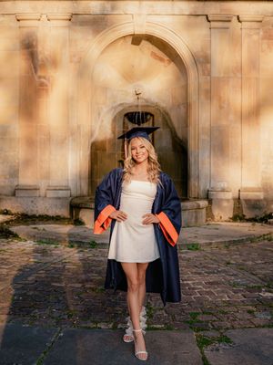 Graduate wearing navy blue and orange academic regalia poses under stone archway on graduation day.