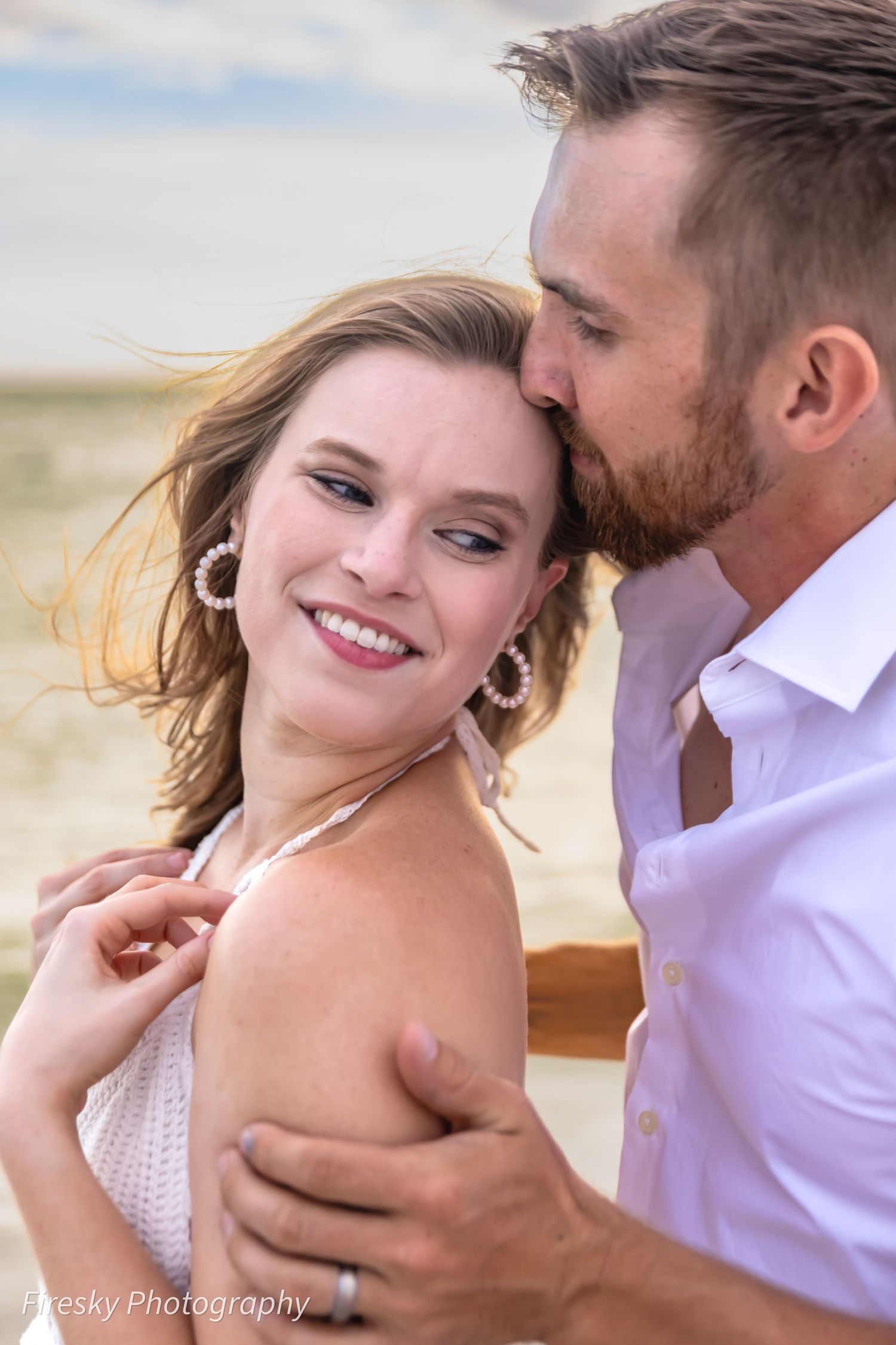 A romantic couple embraces outdoors with wind-blown hair as sunlight illuminates their joyful moment together.