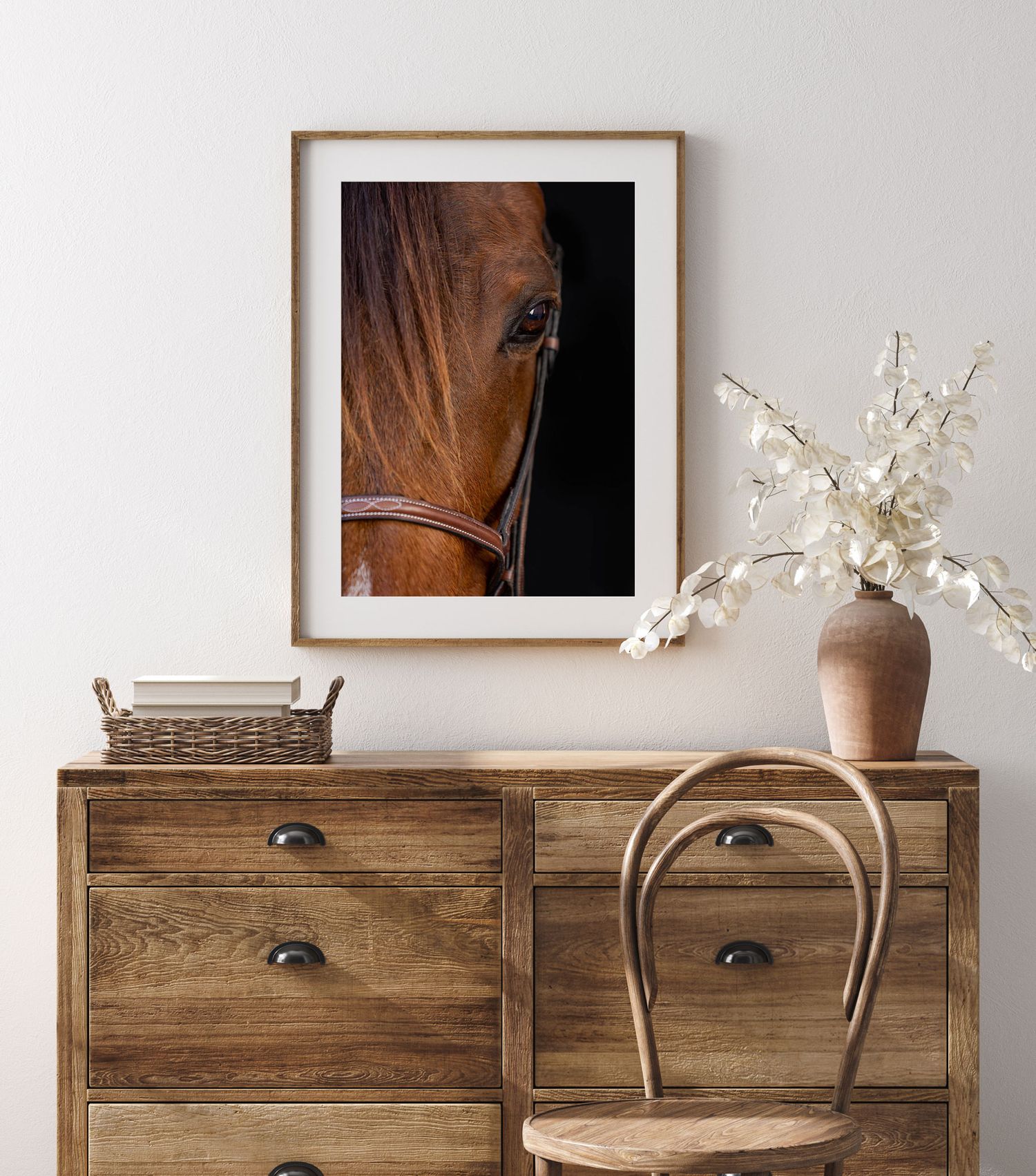 A framed horse portrait above rustic wooden dresser with vintage chair and white floral arrangement in earthen vase.