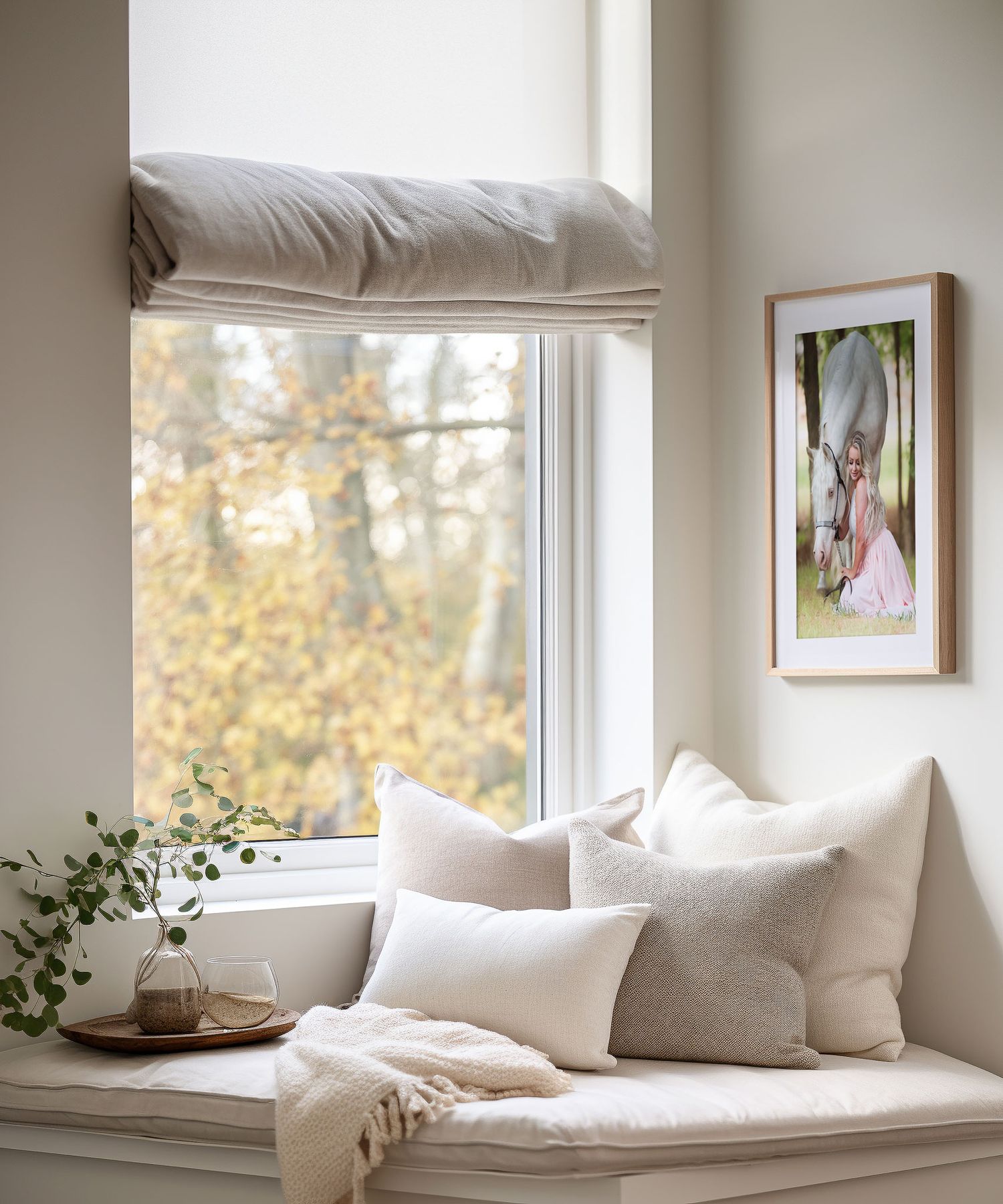 Cozy window seat nook with white cushions, throw pillows and a framed photo of a woman and her horse on wall beside autumn trees outside.