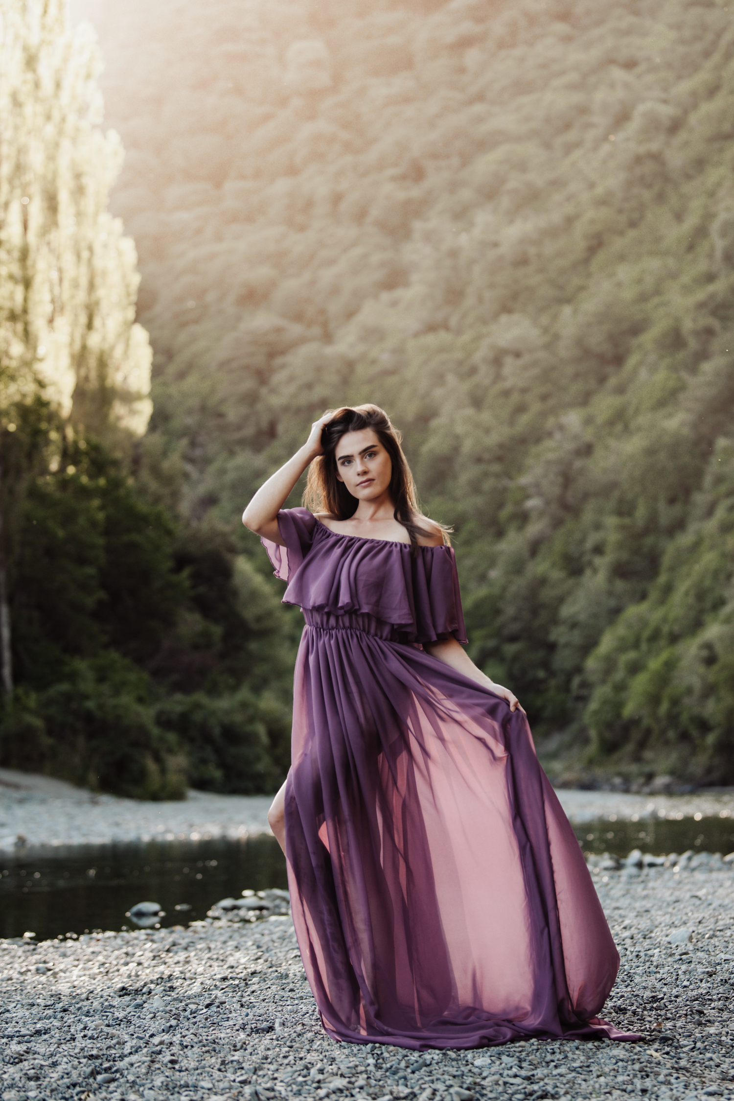 A flowing purple off-shoulder maxi dress billows in the breeze by a riverbank with forested hills in the background.