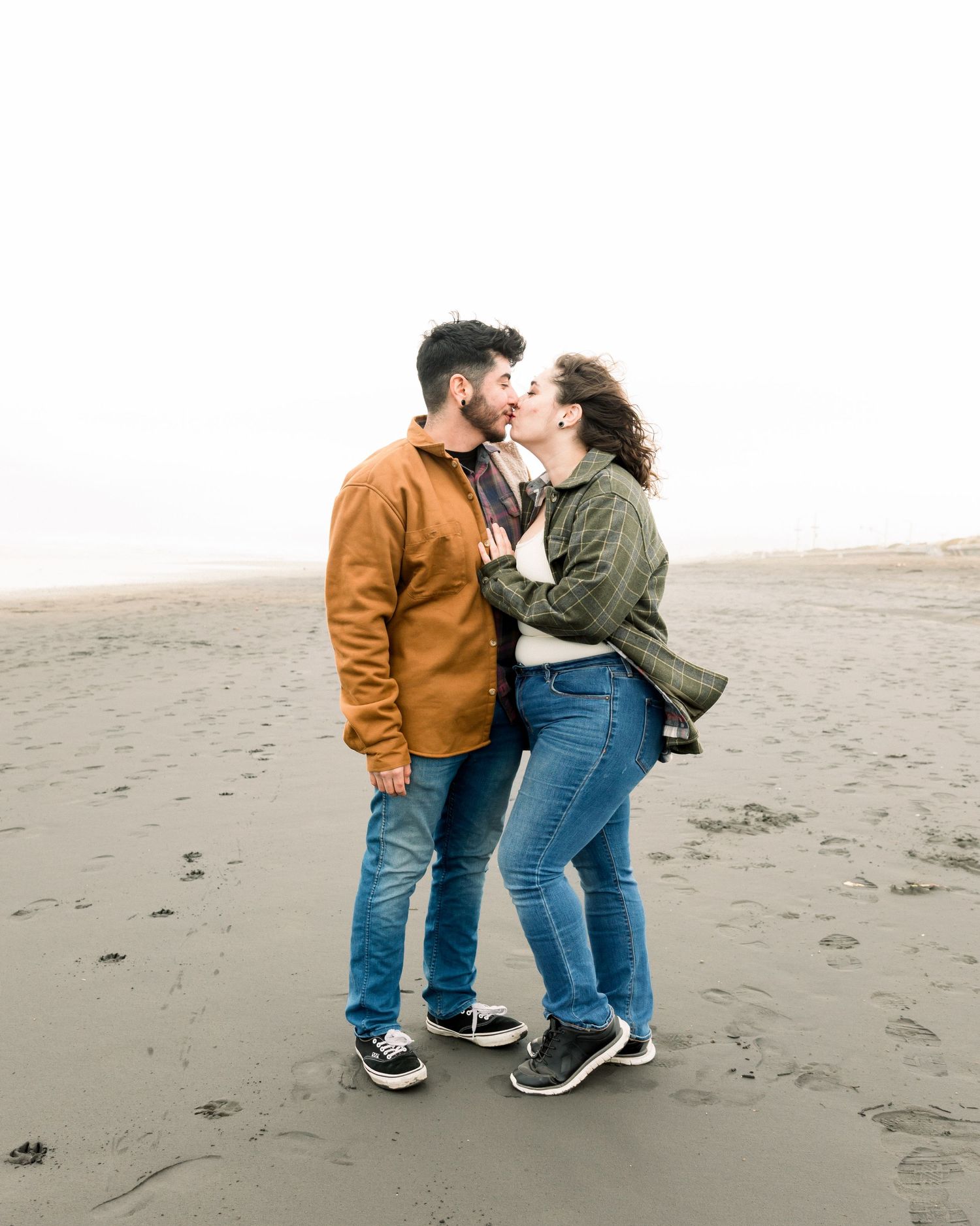 Couple embracing on foggy beach wearing casual denim and jackets.