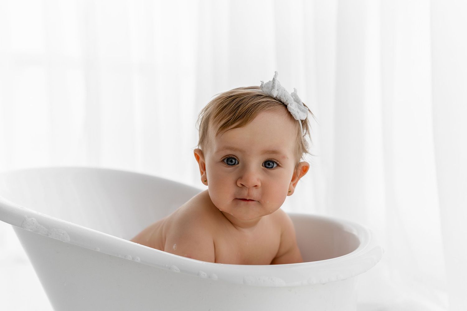 Baby with bow sitting in elegant white bathtub against light backdrop.