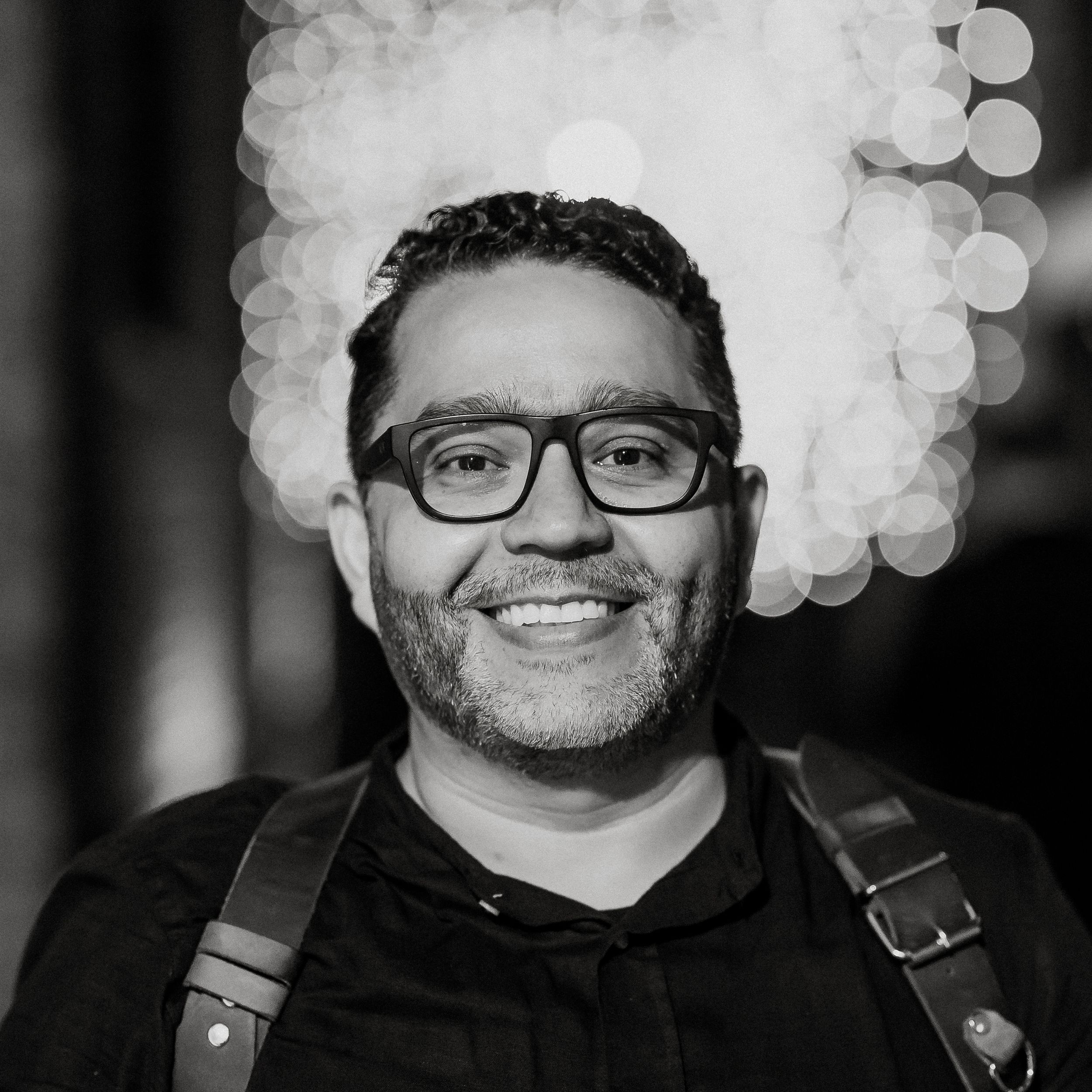 Black and white portrait of smiling male photographer with glasses and camera strap, bokeh background.