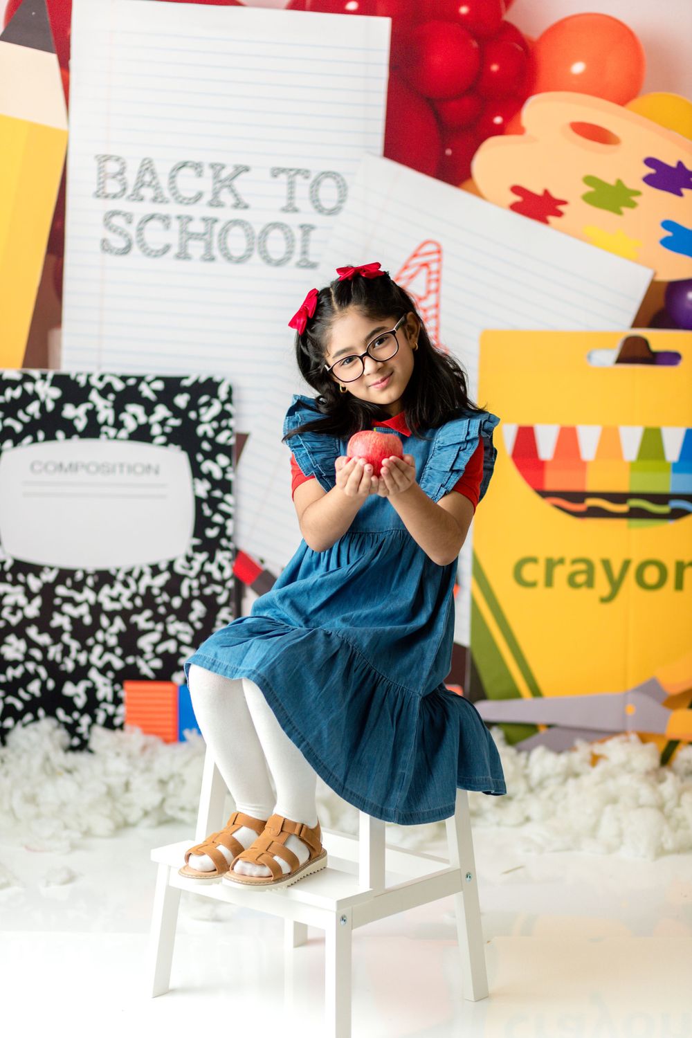 Student in blue dress holding apple on white chair with school supplies and decorative background.