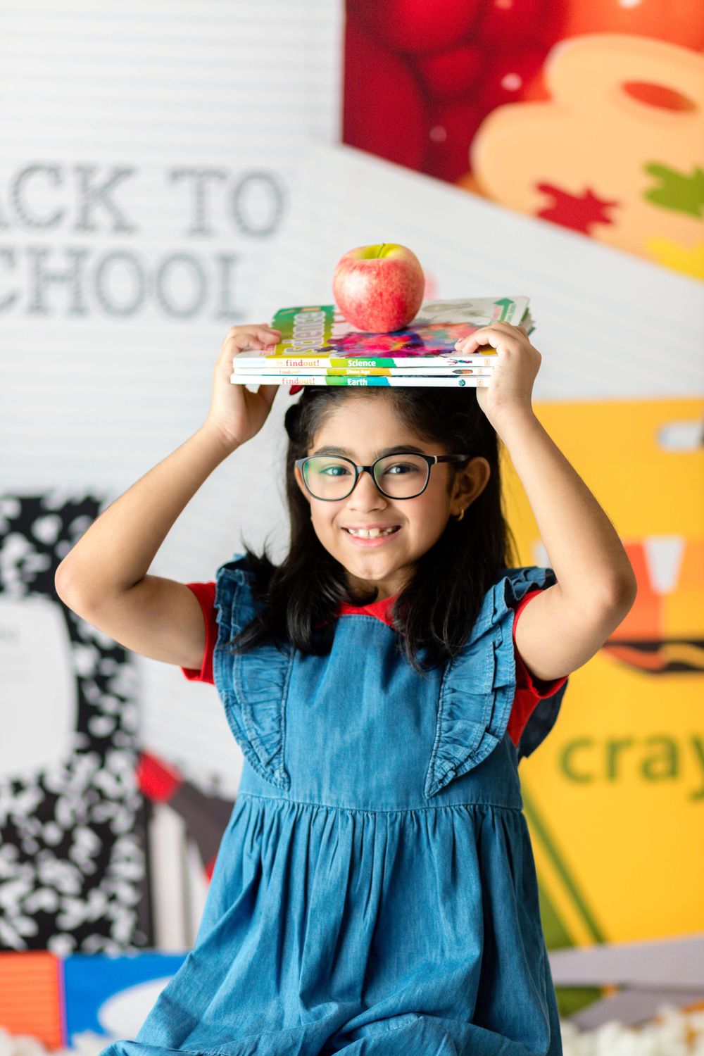 A student balances school supplies and an apple on their head against a colorful back to school backdrop.