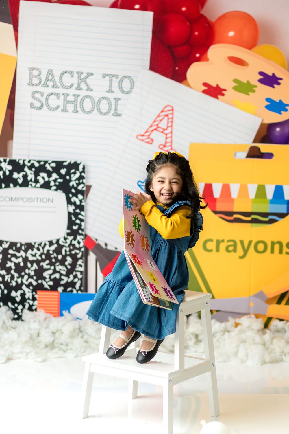 Child in blue dress sitting on chair with back-to-school themed decorations and balloons.